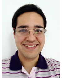 A figura mostra o rosto do professor Adelcio Camilo Machado. Homem branco, de óculos, com cabelos e olhos escuros, sorrindo, vestindo uma camisa listrada nas cores branco e lilás. 