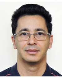 Homem, moreno claro, cabelos curtos pretos, olhos castanhos escuros, com óculos. Face com discreto sorriso. Usa camisa polo azul com listras vermelhas.