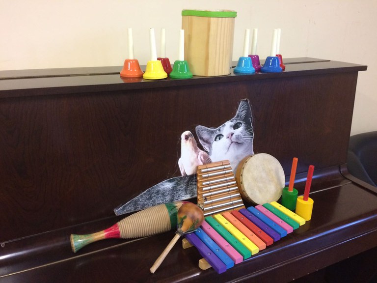 A foto mostra um piano fechado, decorado com a imagem de um gato e um passarinho. Na frente dessa imagem sobre seu tampo, no centro, estão sobrepostos instrumentos de pequena percussão como xilofone, metalofone, 1 tambor de mão, alguns chocalhos e na parte superior há um pequeno tambor e um ogo de sinos de mãos coloridos.